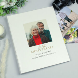 A personalised, cream-coloured photo album commemorating a 50th Golden Wedding Anniversary. The cover features a square photo of an older, smiling couple (Julie and Thomas) centred above the text "50th ANNIVERSARY" in gold script, followed by "JULIE & THOMAS" and "Golden Wedding Anniversary" in black text. The album is displayed on a light gray surface next to a white candle, green foliage, a black Nikon camera lens, and several loose, candid family photos.
