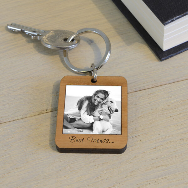 A rectangular solid cherry wood keyring cut into a classic Polaroid style frame, featuring a printed black and white photo of a smiling girl hugging her white dog. Engraved text below the photo reads "Best Friends....". The keyring sits on a light wooden desk next to a silver key and a black book.