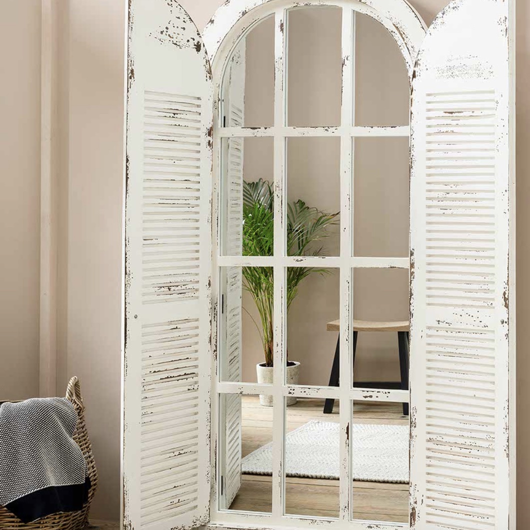 The white arched window mirror with open shutter doors leaning against a soft beige wall next to a palm plant.