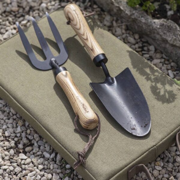 A matching hand garden fork and trowel with blackened steel heads and light ash wood handles resting on a green fabric kneeling cushion.