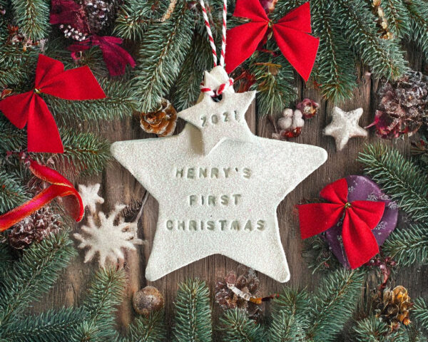 A handcrafted white clay double star Christmas tree decoration hand-stamped with "HENRY'S FIRST CHRISTMAS" and "2021", hanging from red and white twine amidst pine branches and red bows.