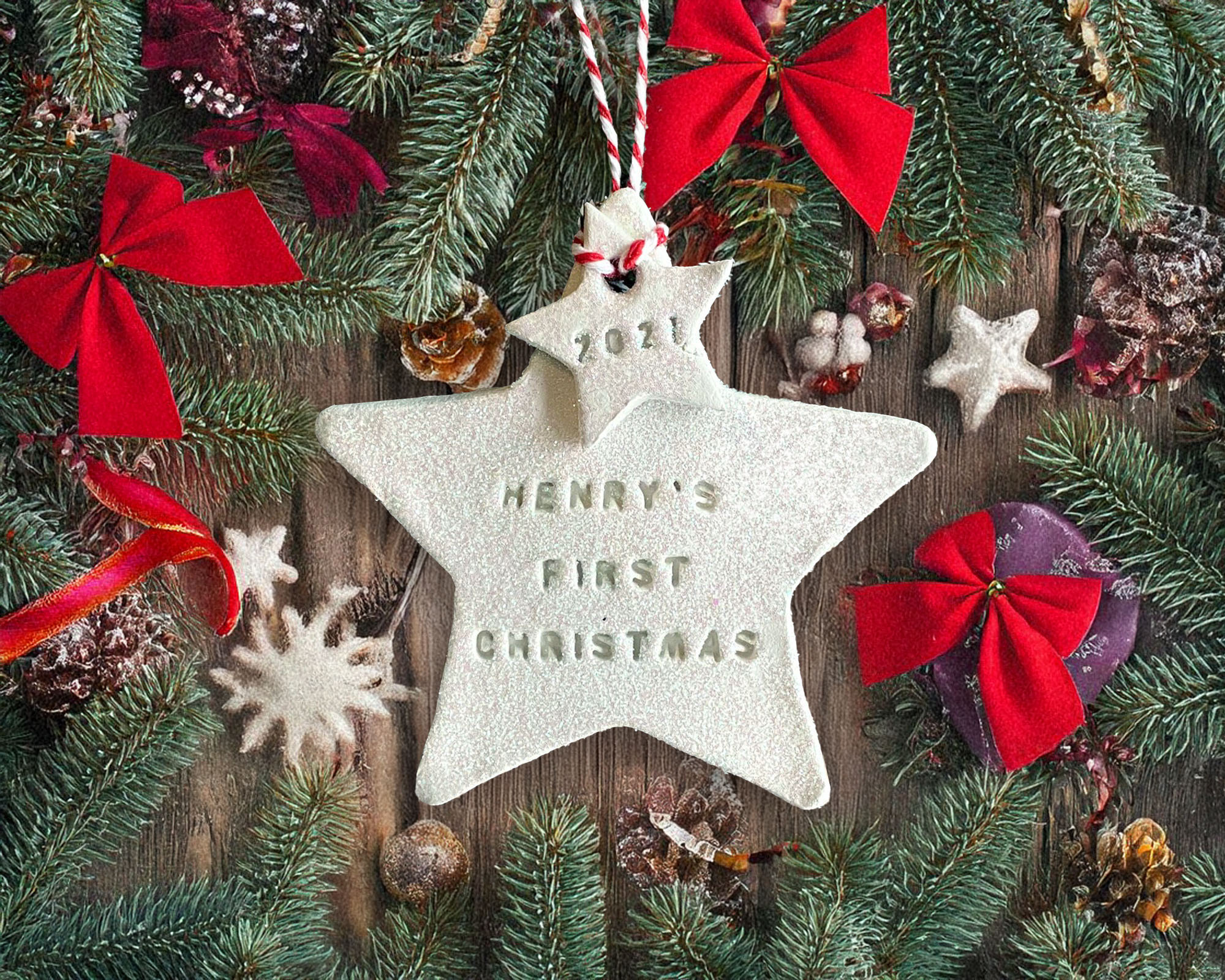 A handcrafted white clay double star Christmas tree decoration hand-stamped with "HENRY'S FIRST CHRISTMAS" and "2021", hanging from red and white twine amidst pine branches and red bows.