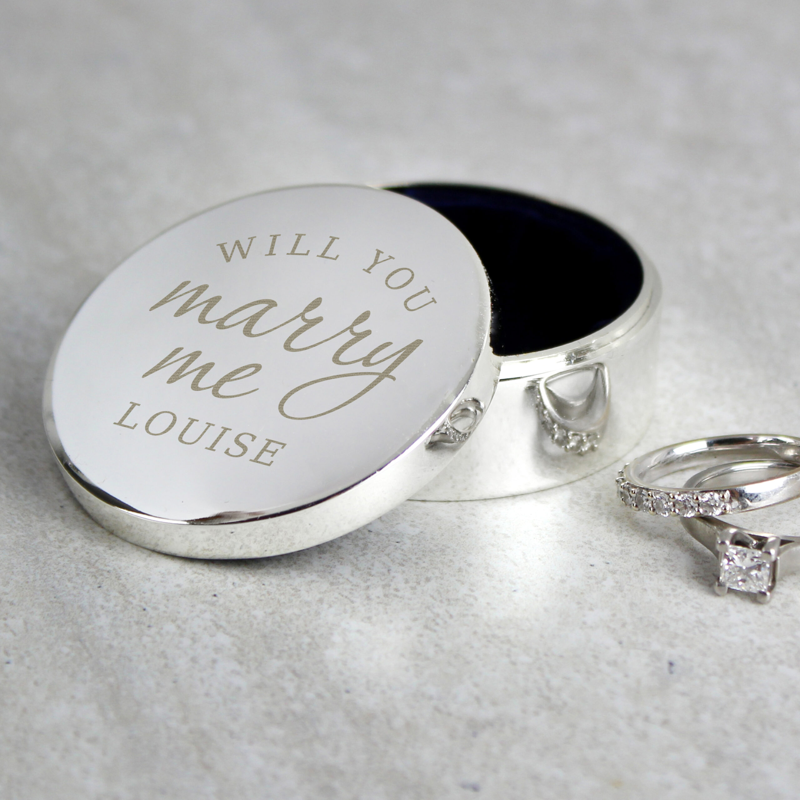 A polished silver round ring box with an open lid engraved with 'WILL YOU marry me LOUISE', styled on a light stone countertop next to a diamond engagement ring and wedding band.