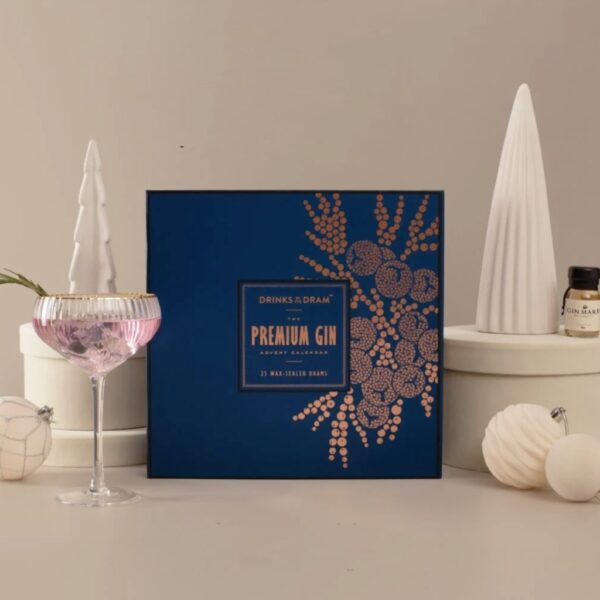 Navy and copper gin advent calendar box containing 25 wax-sealed drams, shown with a pink gin coupe cocktail, a sample dram bottle and cream Christmas decorations