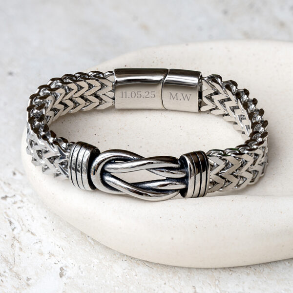 Personalised men’s woven infinity knot bracelet in silver tone with clasp engraved with a date and initials, resting on a cream stone dish