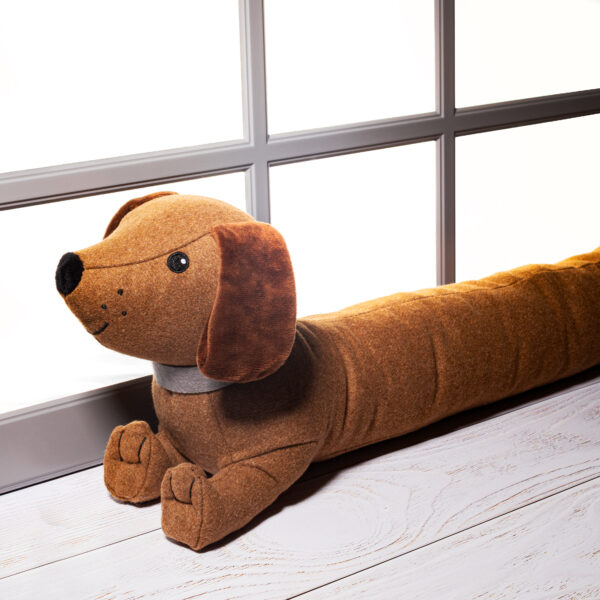 A long, plush, brown tweed-texture sausage dog (Dachshund) draught excluder lying down in front of a window or door frame to block drafts. The dog has soft floppy ears, a stitched nose, and is wearing a simple grey fabric collar.