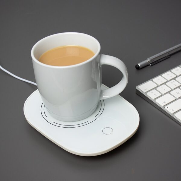 A white oval USB cup warmer sitting on a grey desk next to a computer keyboard and a pen, with a white mug of tea resting on the warming plate.