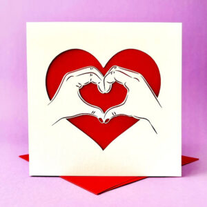 Square white papercut card featuring a laser-cut design of two hands forming a heart shape, set against a vibrant red heart background and displayed with a matching red envelope.