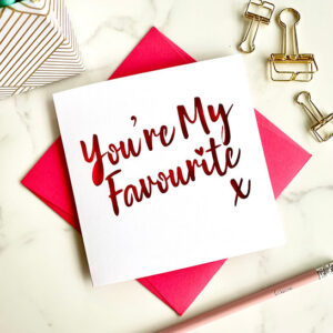 Square white papercut card featuring the laser-cut words "You're My Favourite" in a script font with a small heart, layered over a vibrant pink background and styled on a marble surface with gold office accessories.