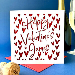 Square white papercut card featuring dozens of laser-cut hearts and the text "Happy Valentine's James" in a cursive font, layered over a red background and displayed with a red envelope and champagne bottle.