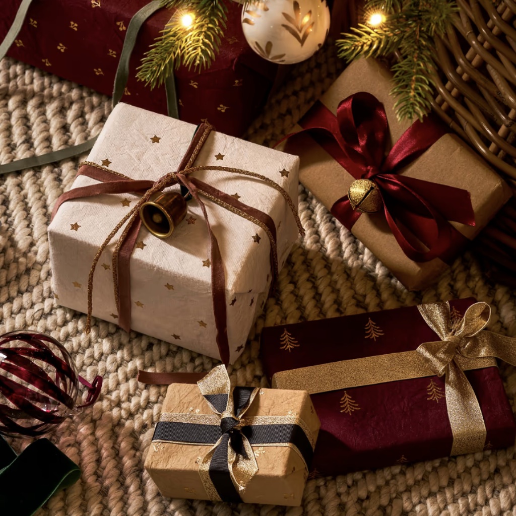 Selection of Christmas presents wrapped in berry red, warm white, and truffle cotton wrapping paper decorated with gold foil trees and stars.