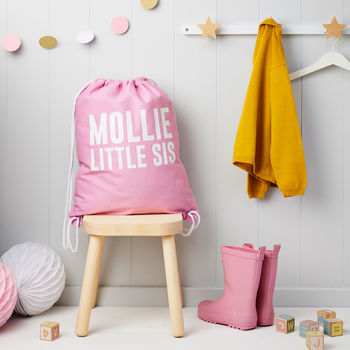 Girl's personalised pink gym drawstring bag printed in white with MOLLIE LITTLE SIS, sitting on a wooden stool.