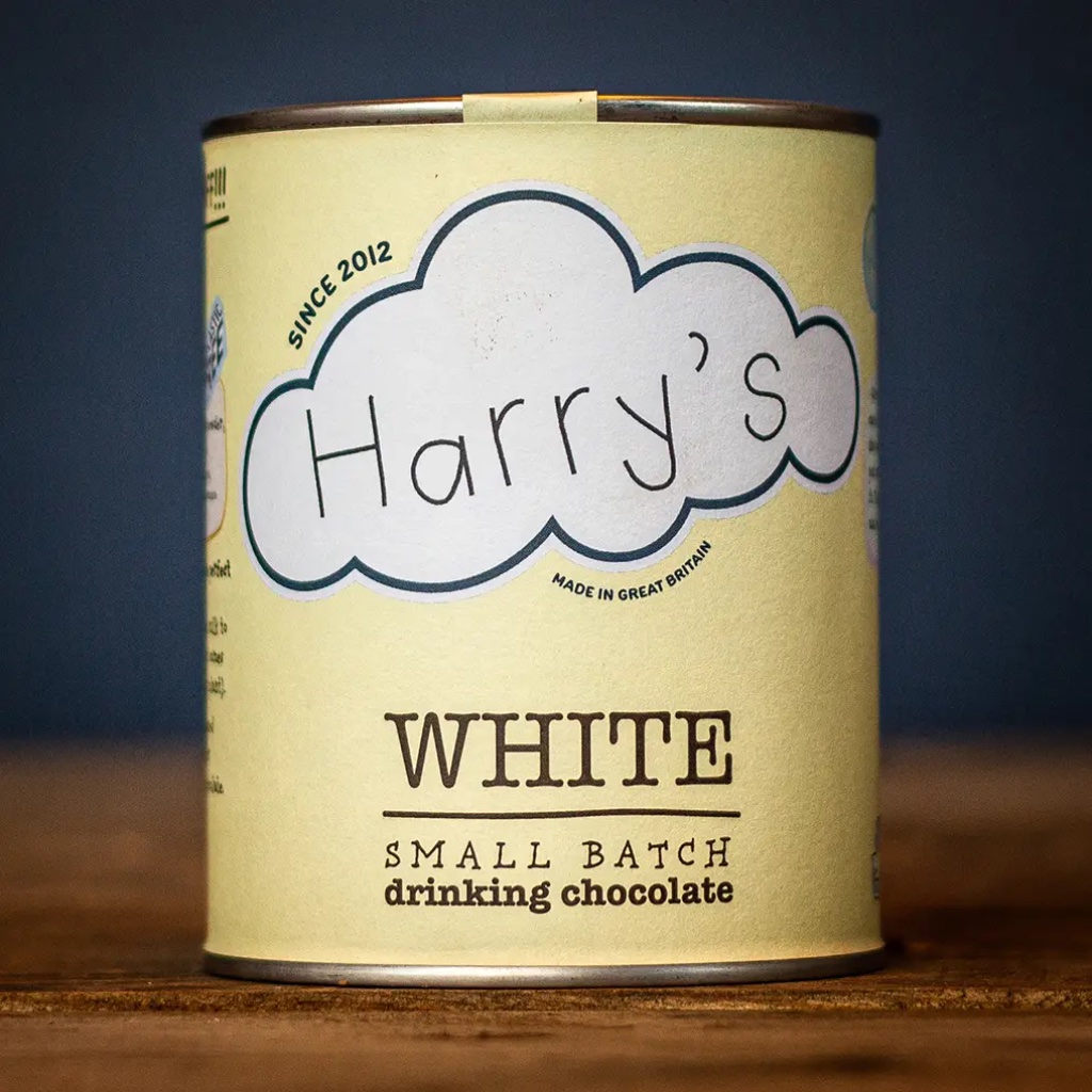 Recyclable soft yellow tin of Harry's Small Batch White Drinking Chocolate resting on a rustic wooden table.