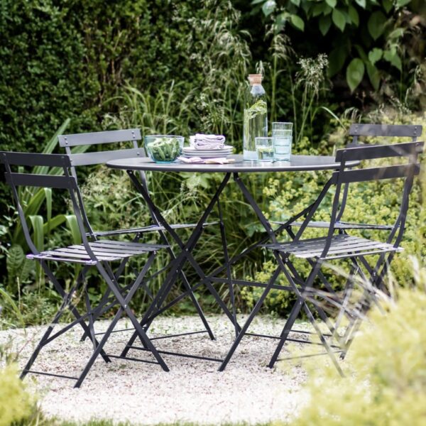 A French-inspired carbon dark grey metal bistro set featuring a round dining table and four folding chairs on a gravel patio surrounded by lush green foliage.