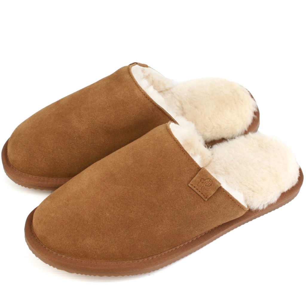 A pair of men's slip-on slipper mules showing the rich chestnut tan suede exterior, a thick trim of natural cream sheepskin wool at the opening, and a subtle matching tan pull-tab on a white background.