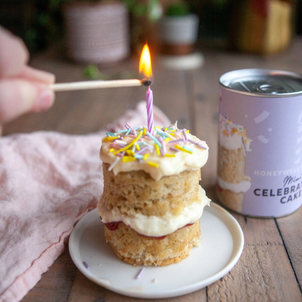 A hand using a wooden match to light a candle on a mini layered celebration cake decorated with icing and colourful sprinkles, placed next to a Honeywell Bakes Mini Celebration Cake tin.