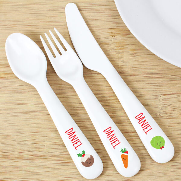 A white plastic children's cutlery set personalised with the name 'DANIEL' and decorated with cute Christmas food motifs, lying on a wooden tabletop next to a white plate.