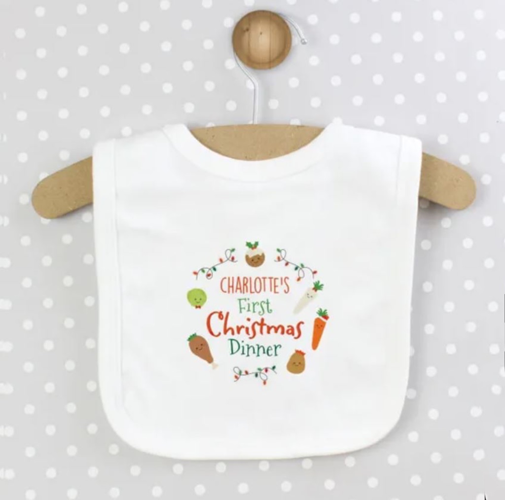A white cotton baby bib custom printed with 'CHARLOTTE'S First Christmas Dinner' surrounded by illustrated festive food characters, hanging on a wooden hanger against a grey polka dot background.