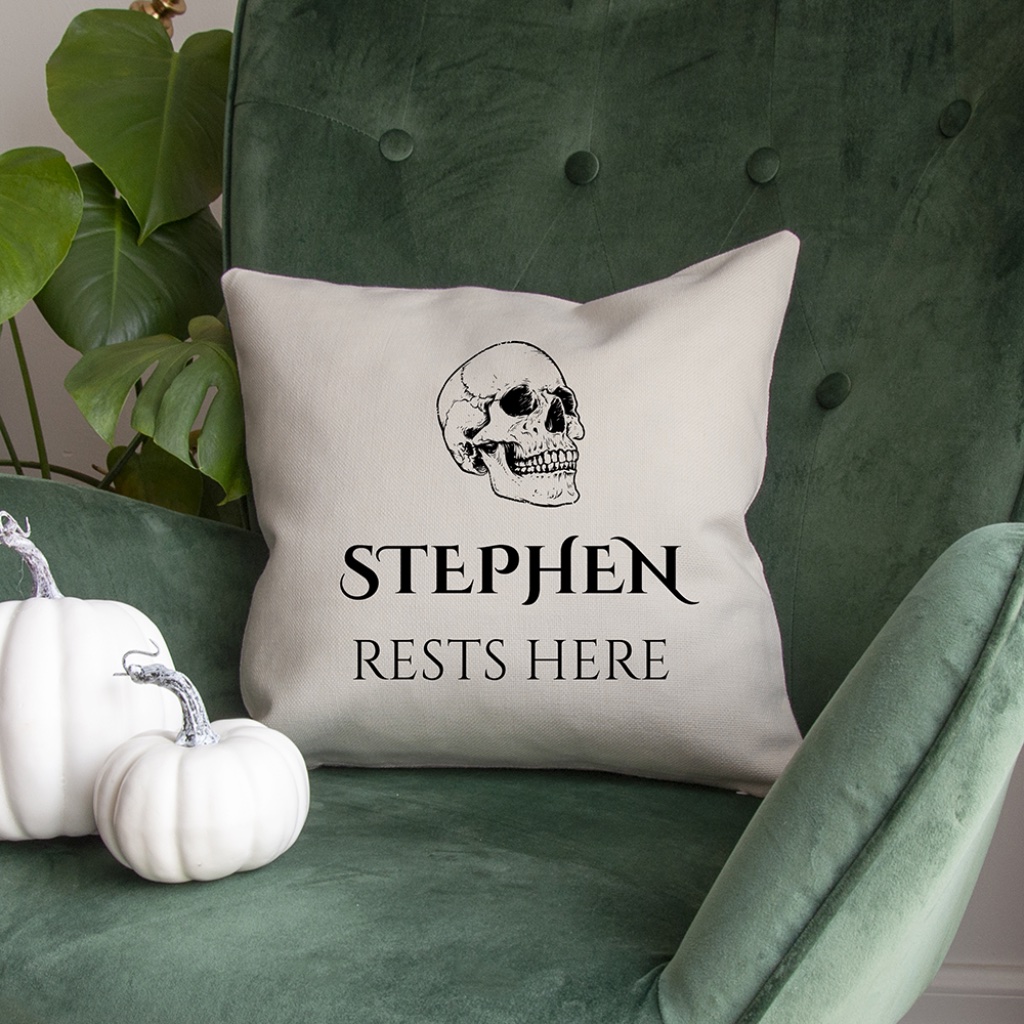 Square white cushion cover with skull graphic and Stephen Rests Here text resting on a dark green velvet armchair next to decorative white pumpkins