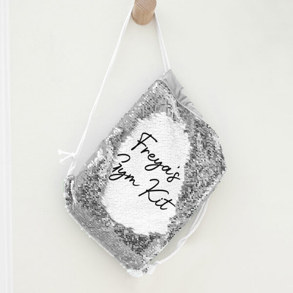 Personalised kids silver sequin drawstring bag hanging on a wooden wall peg, showing the custom revealed text reading Freya's Gym Kit.