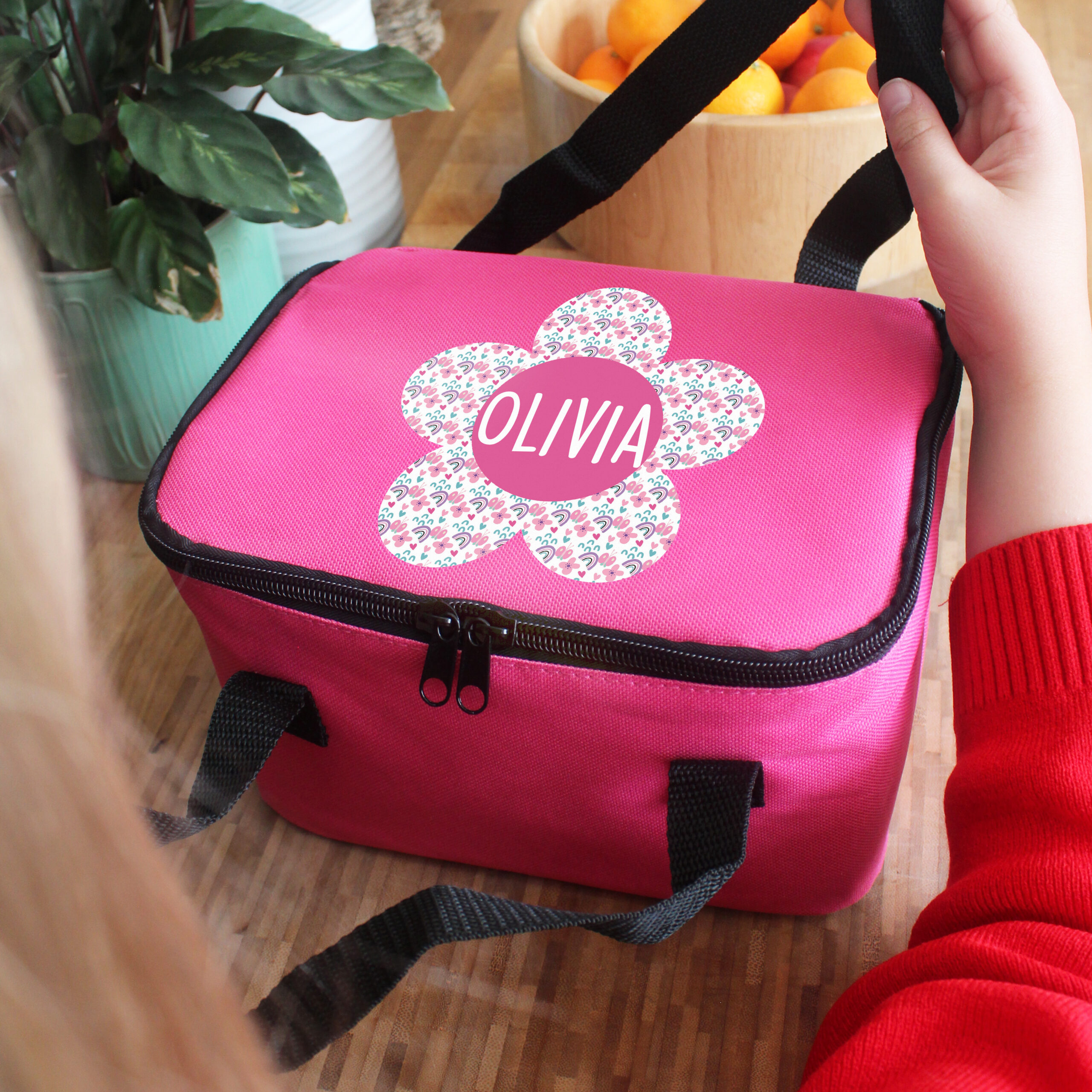 An insulated pink children's lunch box bag custom printed with the name Olivia, featuring a large patterned flower design with rainbows, hearts, and stars on a wooden surface.