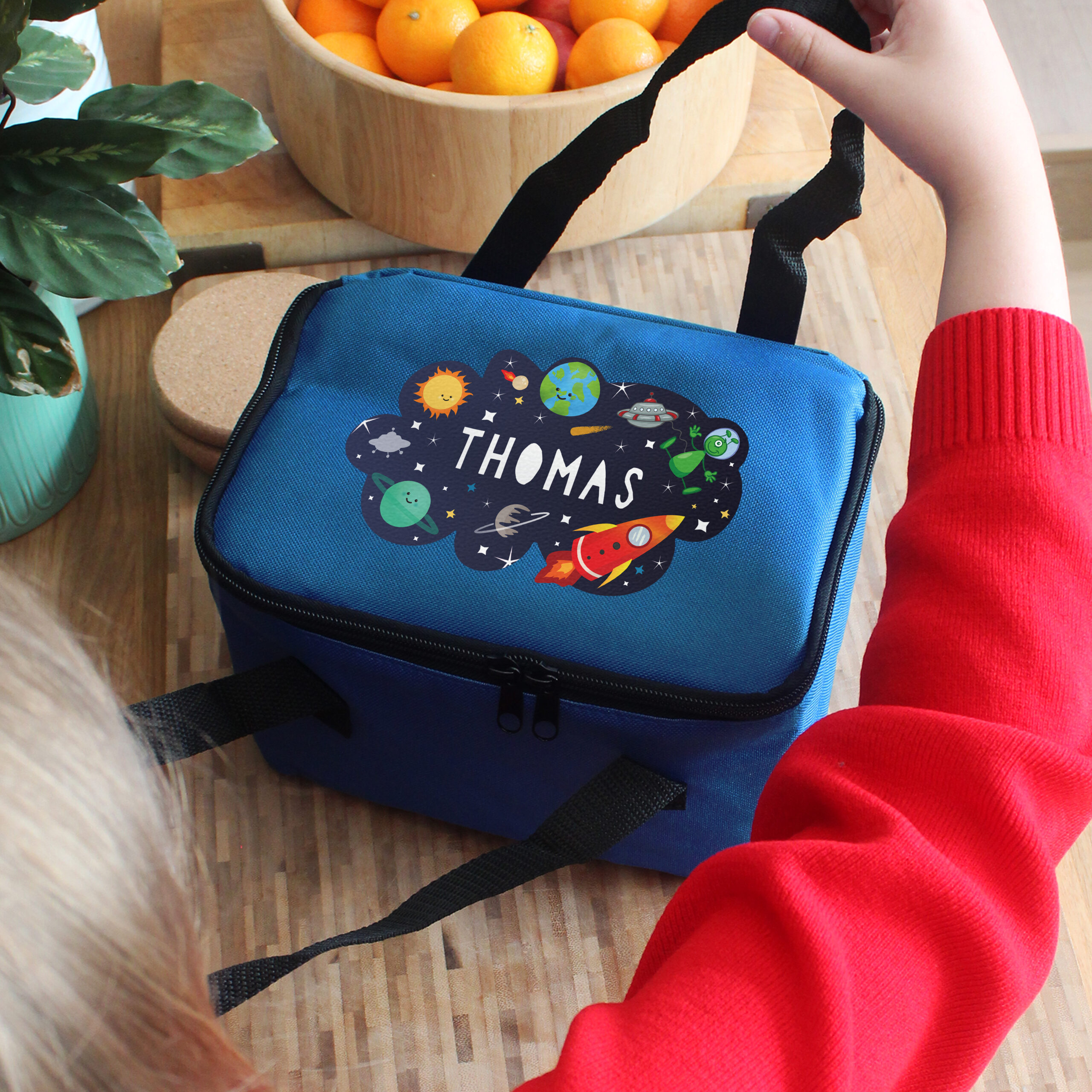 A child wearing a red jumper holds the strap of a personalised blue space lunch bag printed with the name Thomas.