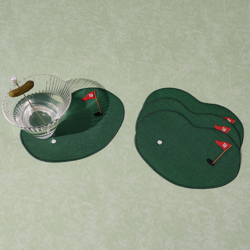 A set of four green linen cocktail napkins shaped like golf putting greens with embroidered white balls and red 19 flags, featuring a crystal cocktail glass sitting on one napkin.