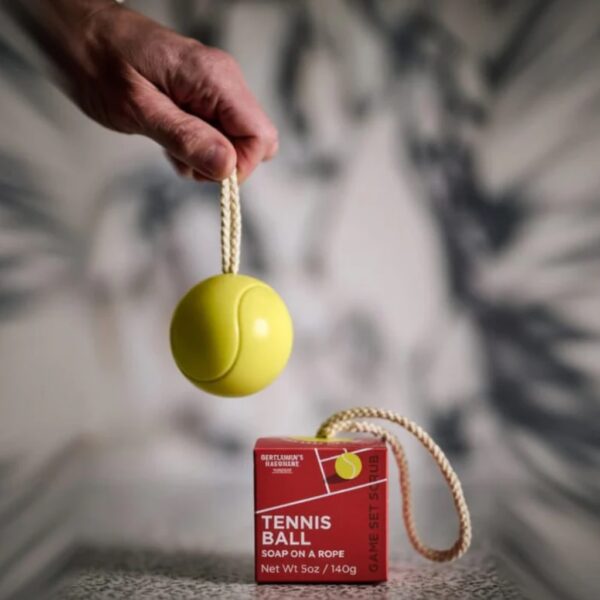 A hand holding up a yellow tennis ball shaped soap on a woven rope next to its red court-themed gift packaging.