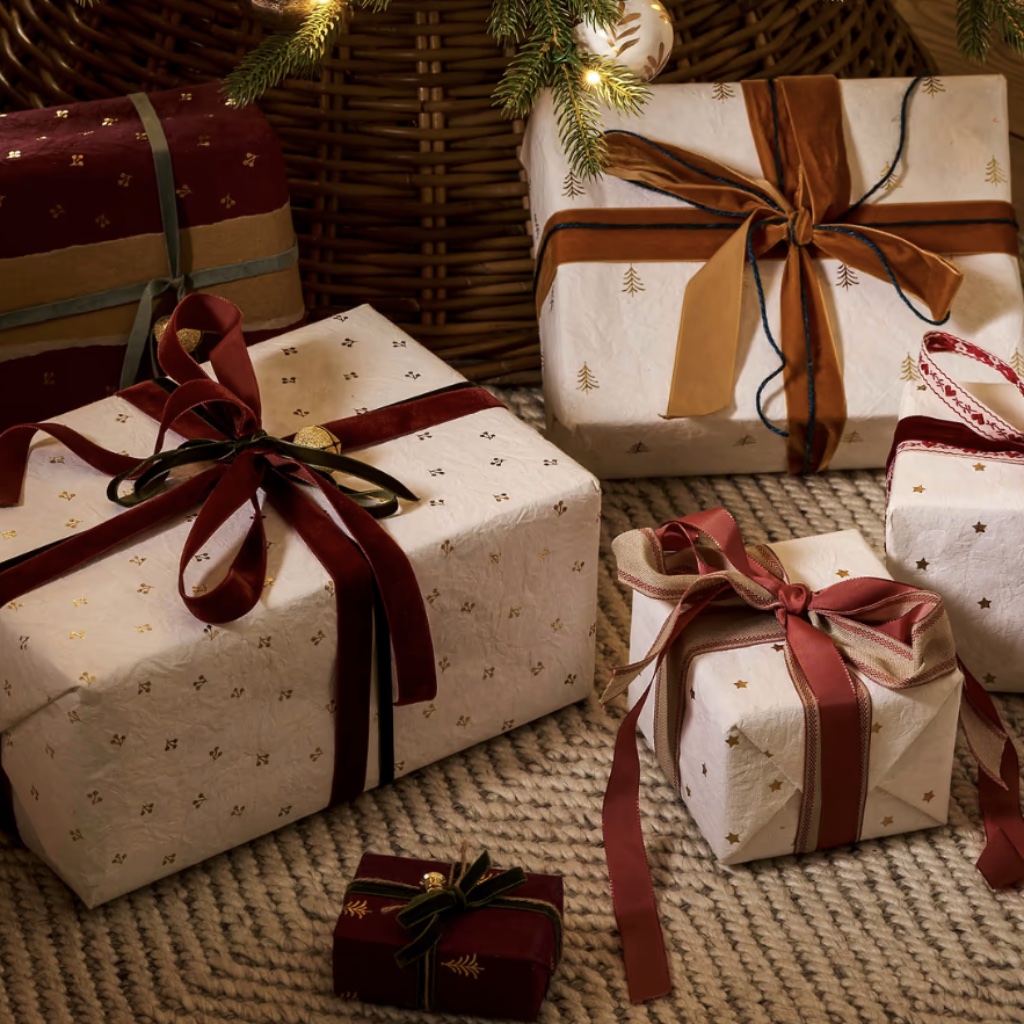 Selection of Christmas gifts beautifully wrapped in warm white handmade cotton paper with gold foil trees, stars, and berries, finished with velvet ribbons under a tree.