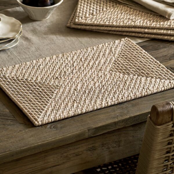 Handwoven white washed rectangular rattan placemat styled on a rustic wooden dining table alongside silverware and napkins.