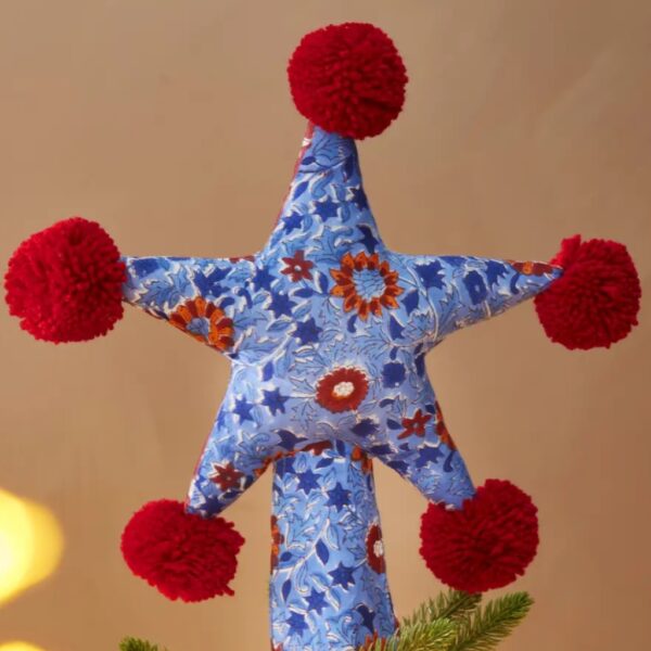 Blue floral block print star tree topper with five red pom poms on top of a lit Christmas tree