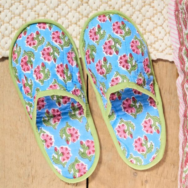 Pair of blue floral block print cotton slippers with pink flowers and green piping on a wooden floor beside a woven rug