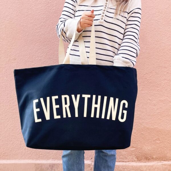 Woman holding an extra large midnight blue canvas tote bag printed with EVERYTHING in off-white letters against a pink wall