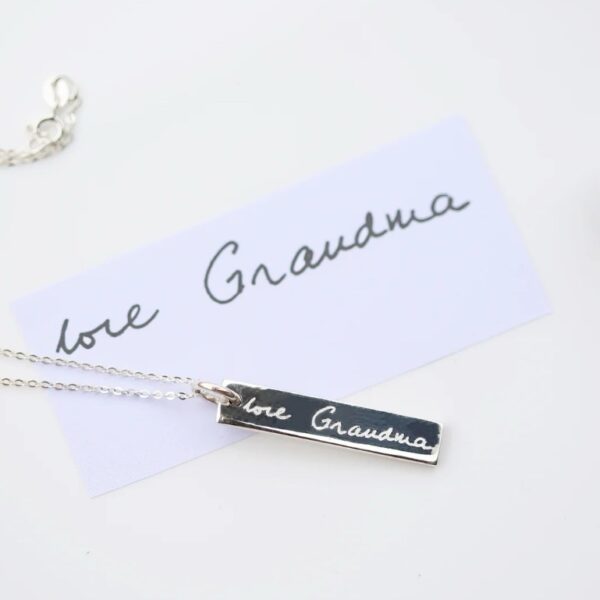 Sterling silver bar necklace engraved in handwriting with love Grandma, lying on the original handwritten note beside a silver pen