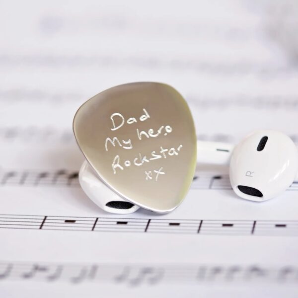 Stainless steel guitar pick engraved in handwriting with Dad My hero Rockstar xx, resting on sheet music beside earbuds