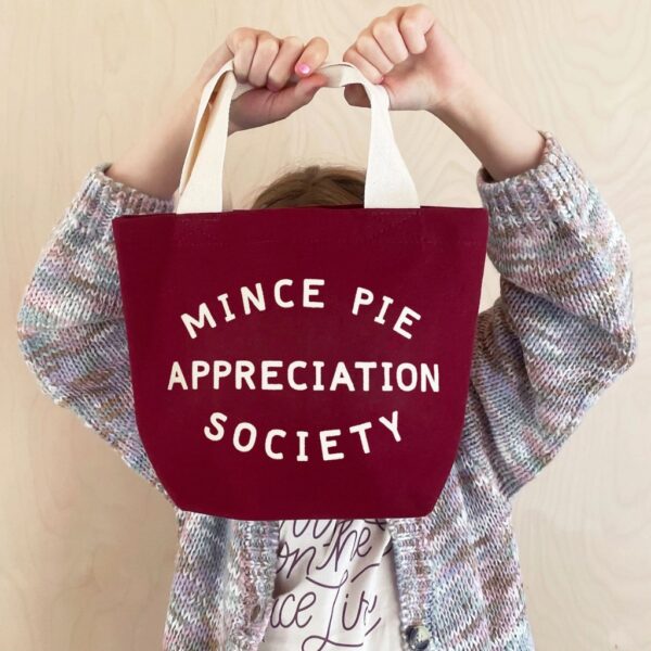 Child holding up a burgundy mini canvas tote bag printed with Mince Pie Appreciation Society in off-white