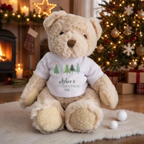 Personalised teddy bear in white T-shirt printed with fir trees and Asher's 1st Christmas 2026 wording, sat by a Christmas tree