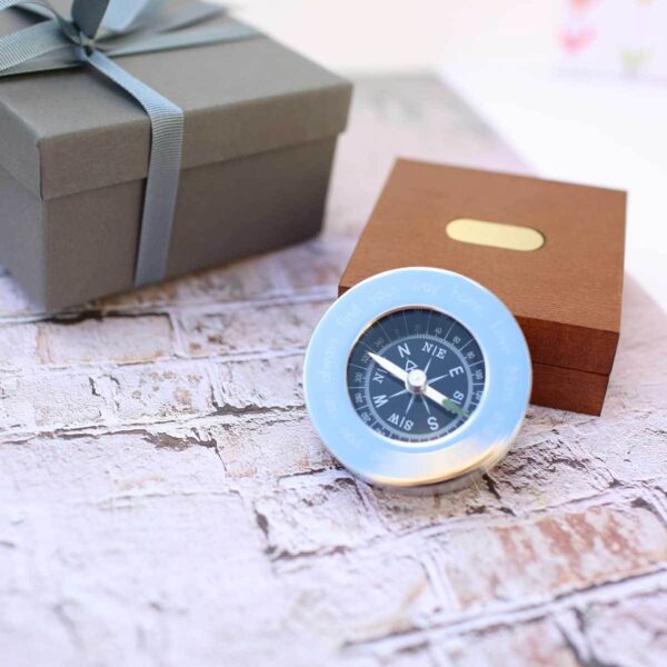 Personalised compass with a black dial and polished rim engraved with a message ending Love Mum, in front of a mahogany box with a brass plate and a grey ribbon-tied gift box
