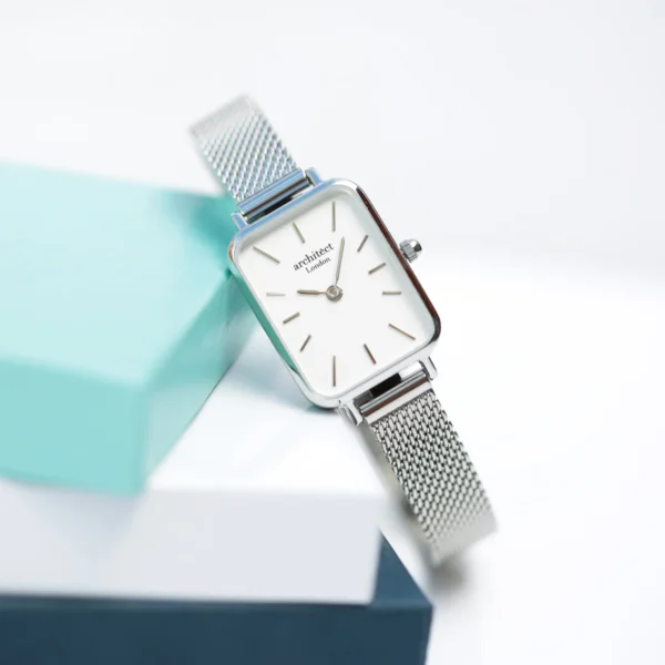 Petite ladies watch with a rectangular white dial, silver baton markers and a polished stainless steel case on a silver mesh strap, resting on stacked gift boxes