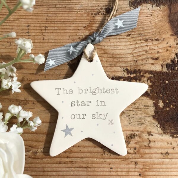 Personalised memorial ceramic star decoration stamped with The Brightest Star In Our Sky tied with a grey star ribbon on rustic wood.
