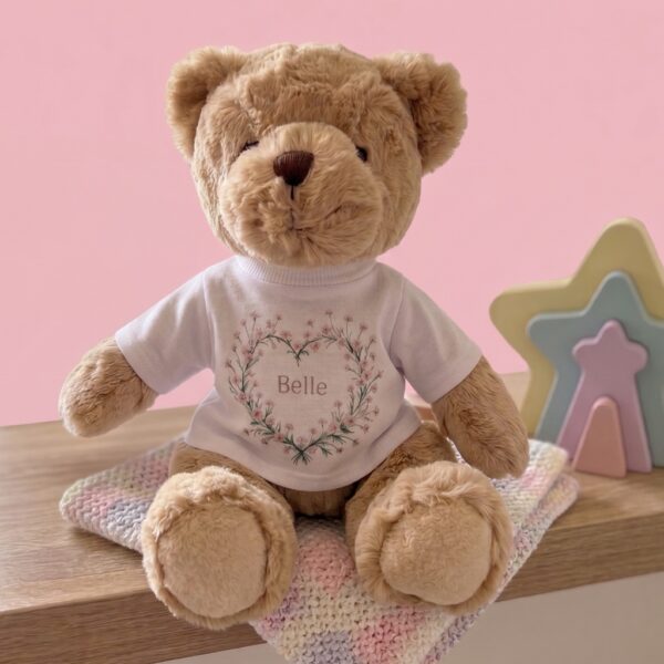 Personalised teddy bear in a pink flower heart T-shirt with the name Belle, sitting on a nursery shelf with a pastel blanket and rainbow star toy