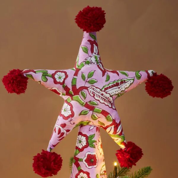 Pink floral block print fabric star tree topper with red pom poms on top of a lit Christmas tree.