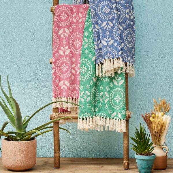 Three recycled cotton jacquard throws in blue, green and red with cream fringing, draped over a bamboo ladder against a turquoise wall