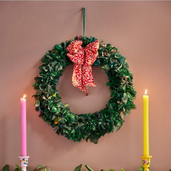 Green recycled sari wreath with red patterned bow hanging above a mantelpiece with pink and yellow candlesticks