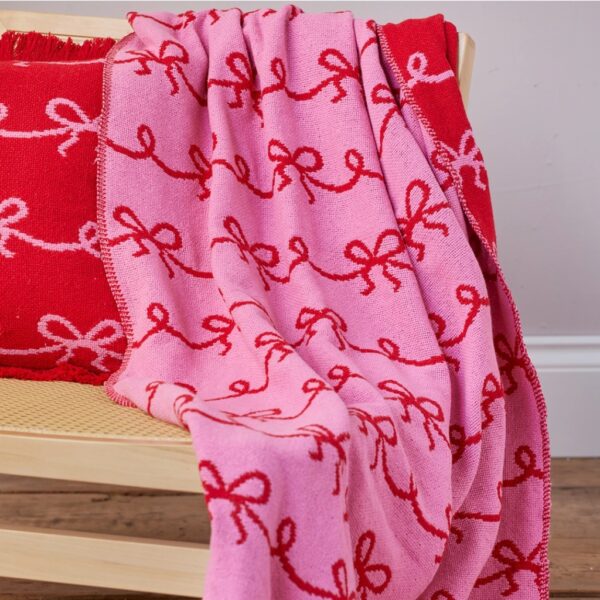 Reversible pink and red bow Christmas throw with fringed edges draped over a wooden bench beside a red bow cushion