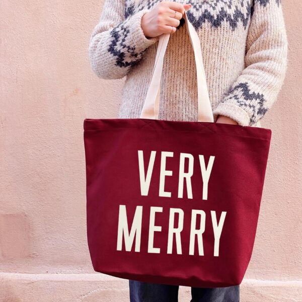 Woman in a Fair Isle jumper holding a burgundy canvas tote bag printed with VERY MERRY in off-white letters
