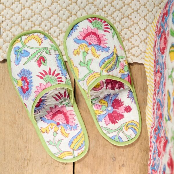 Pair of white floral block print cotton slippers with green piping on a wooden floor beside a woven rug