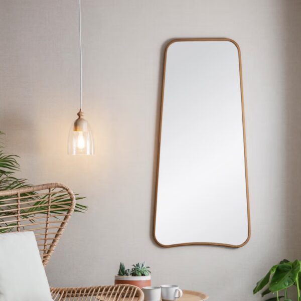 Tall curved bronze wall mirror hanging on a neutral wall beside a pendant light and rattan chair